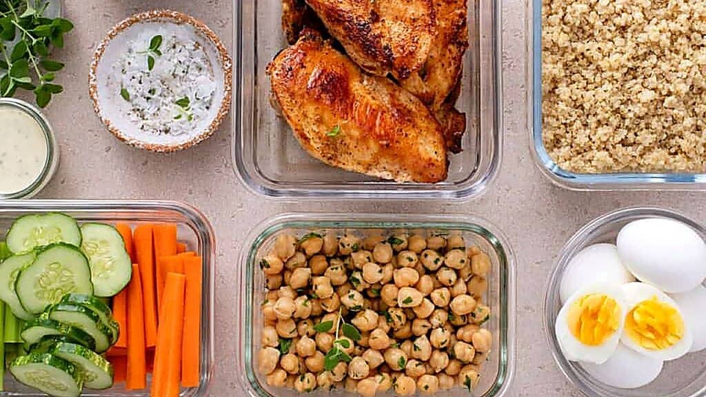 Glass containers with grilled chicken, quinoa, chickpeas, sliced cucumber and carrots, hard-boiled eggs, and a bowl of sauce are arranged on a light surface—perfect for meal prepping to feed your family for less.