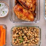 Glass containers with grilled chicken, quinoa, chickpeas, sliced cucumber and carrots, hard-boiled eggs, and a bowl of sauce are arranged on a light surface&mdash;perfect for meal prepping to feed your family for less.