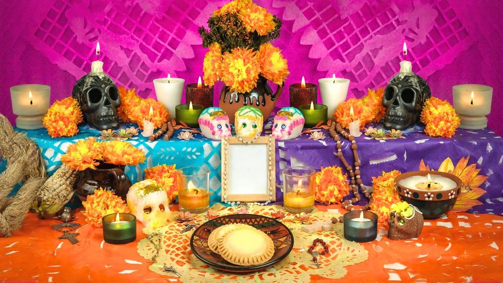 A colorful Day of the Dead altar with marigolds, candles, sugar skulls, decorated paper, food offerings, and a blank photo frame against a vibrant pink and purple background.