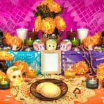 A colorful Day of the Dead altar with marigolds, candles, sugar skulls, decorated paper, food offerings, and a blank photo frame against a vibrant pink and purple background.