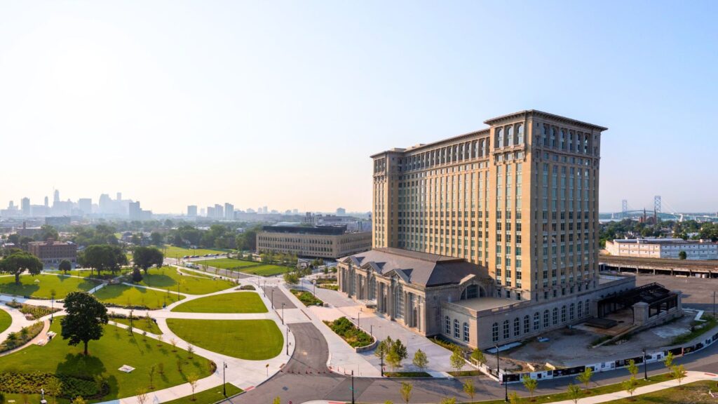 A large historic building with many windows stands next to landscaped green spaces and paved walkways, with a city skyline visible in the background.
