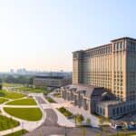 A large historic building with many windows stands next to landscaped green spaces and paved walkways, with a city skyline visible in the background.