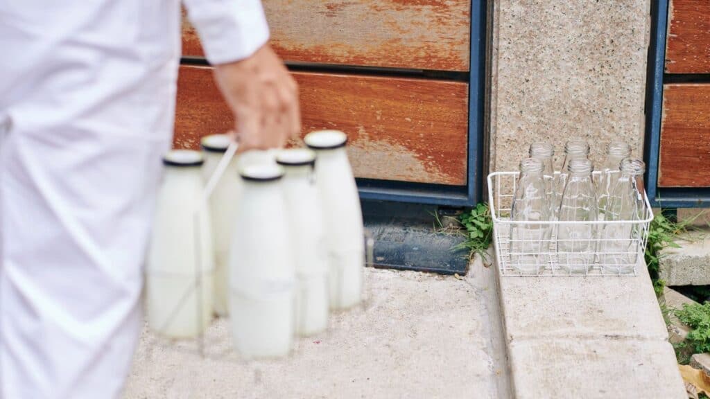 Person in white, evoking classic milkman delivery memories, carries a metal rack of full glass milk bottles toward empty glass bottles in a basket on a doorstep.