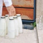 Person in white, evoking classic milkman delivery memories, carries a metal rack of full glass milk bottles toward empty glass bottles in a basket on a doorstep.