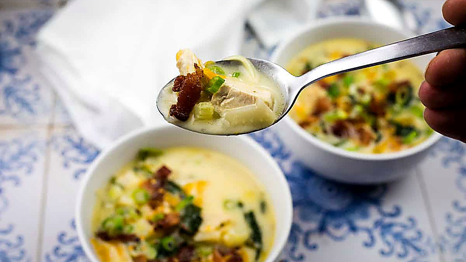 A hand holds a spoonful of Million dollar soup with chicken, bacon, and green onions above a bowl of soup on a blue and white tiled surface.