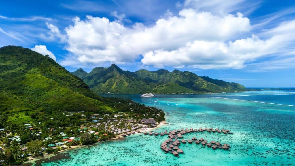 Aerial view of overwater bungalows on a turquoise lagoon, with green mountains, scattered clouds, and a cruise ship in the distance.