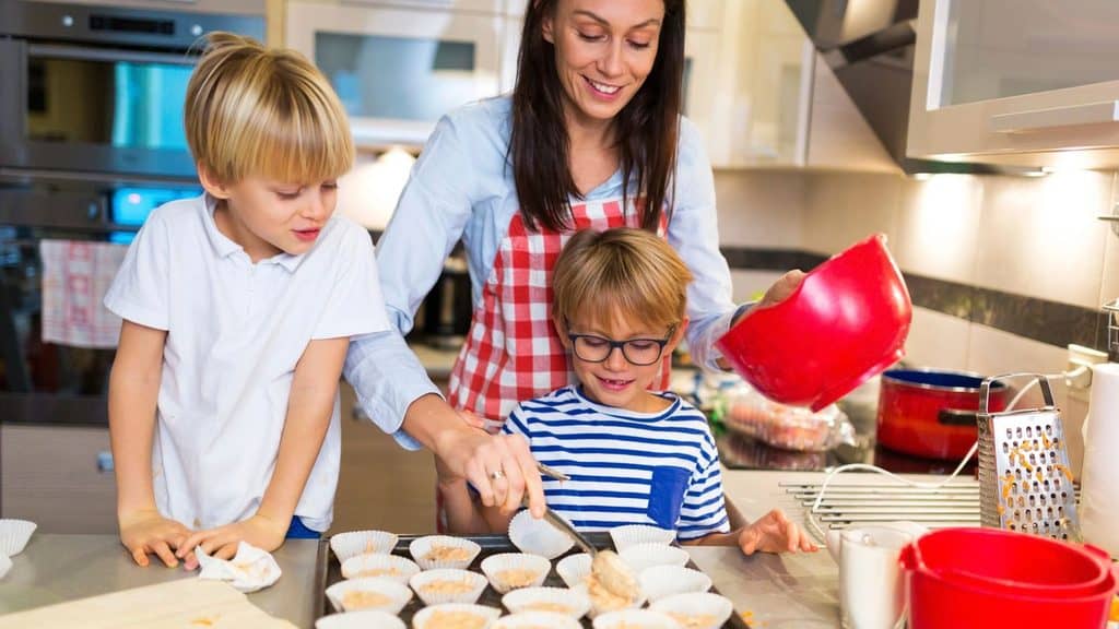 A woman and two young boys prepare muffin batter in a kitchen, using paper liners in a muffin tray. The woman pours batter while one boy fills liners and the other watches.