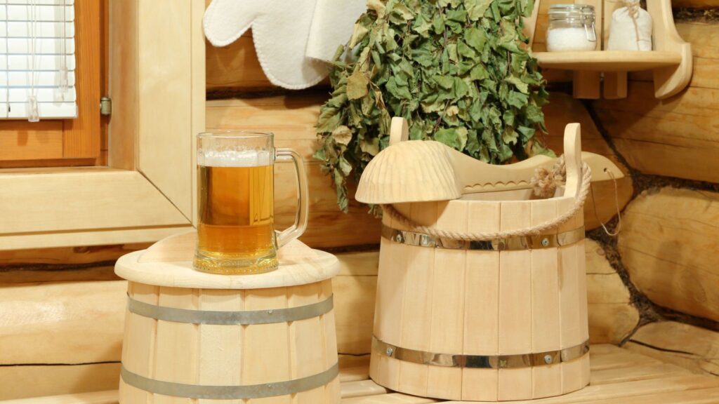 A glass mug of beer sits on a wooden stool next to a sauna bucket, a bundle of leafy branches, and shelves with jars in a wooden interior.