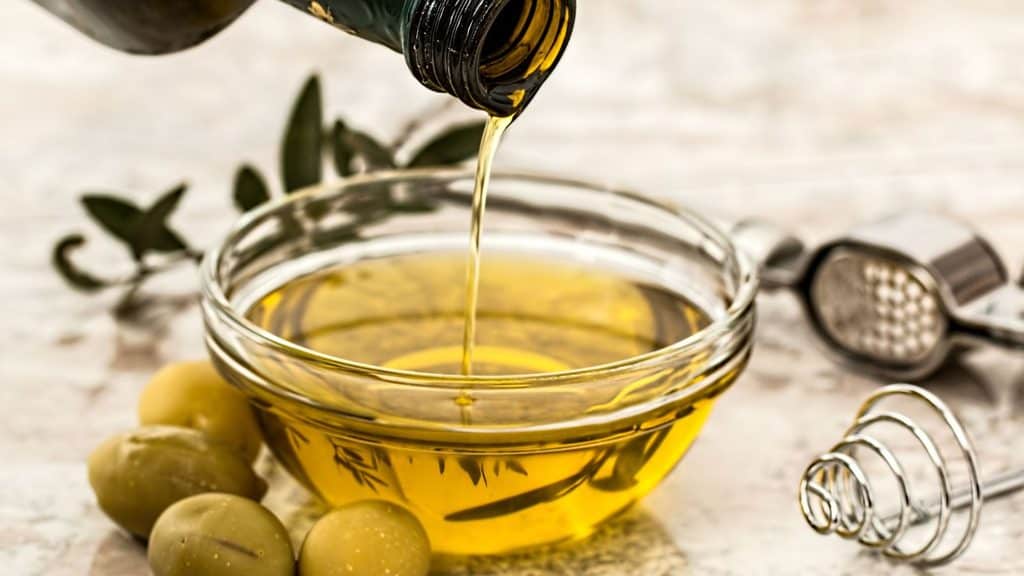 Olive oil is being poured from a bottle into a glass bowl, with whole olives, a whisk, and a garlic press nearby on a light surface&mdash;an inviting scene perfect for celebrating National Extra Virgin Olive Oil Day.