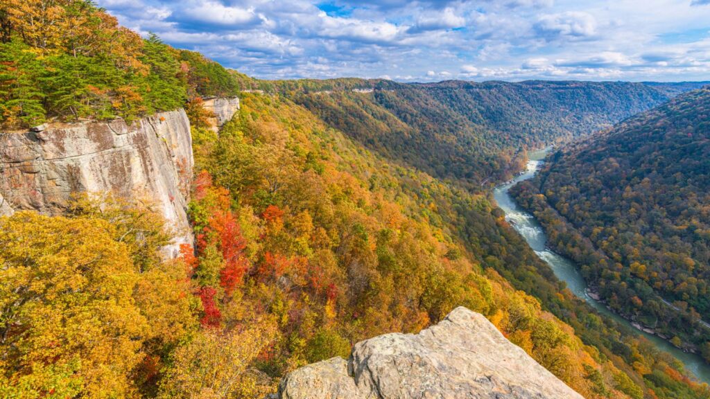 A river winds through a forested canyon with autumn foliage, viewed from a rocky cliff under a partly cloudy sky.