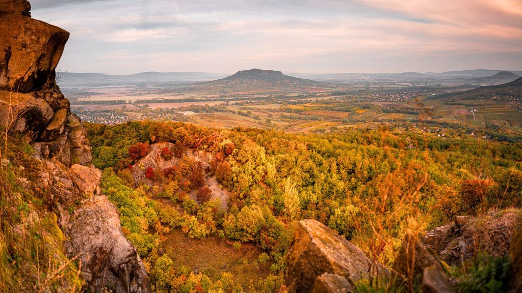 View from a rocky ledge overlooking a colorful autumn forest, with a flat-topped mountain and fields visible in the distance under a partly cloudy sky&mdash;the scene captures the kind of breathtaking beauty that makes autumn the best time to visit Lake Balaton.