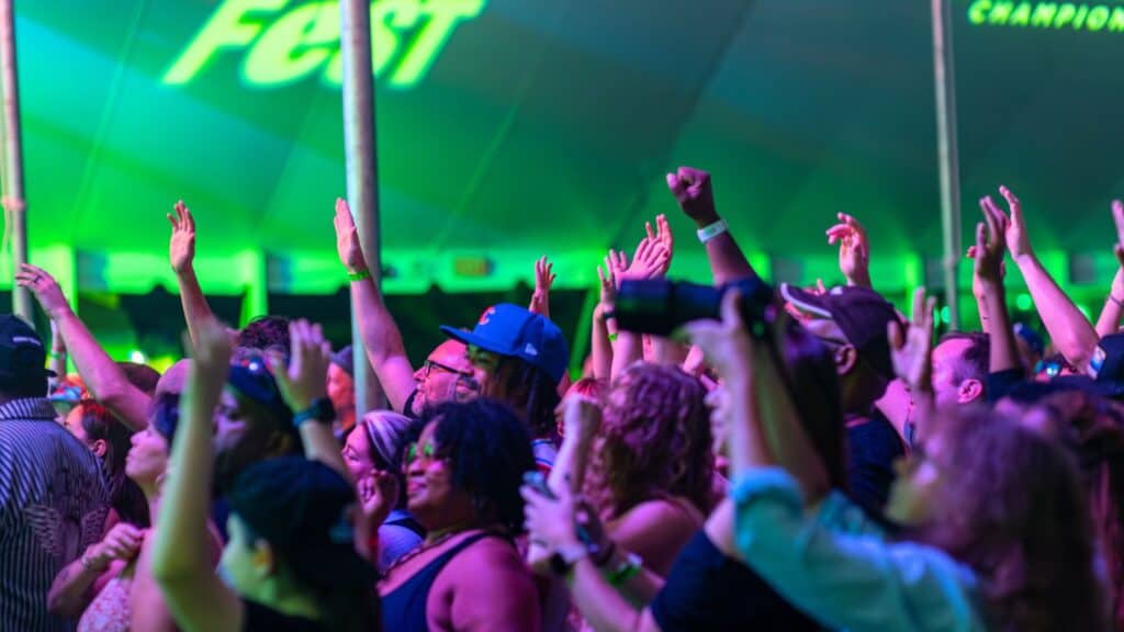 A crowd of people with raised hands enjoy a concert or festival under green lighting inside a large tent.