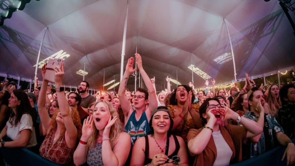 A large crowd of people under a tent cheer and raise their hands at a concert or live event, with some holding phones and others enthusiastically shouting.