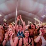 A large crowd of people under a tent cheer and raise their hands at a concert or live event, with some holding phones and others enthusiastically shouting.