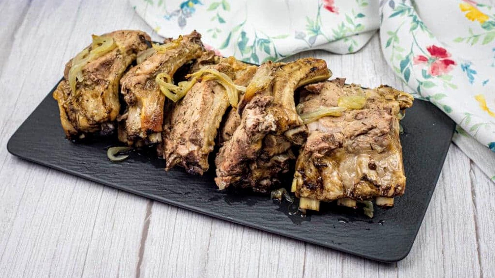 Oven Baked Ribs with cooked onions on a black rectangular slate platter, set on a wooden surface. A floral-patterned cloth is partially visible in the background.