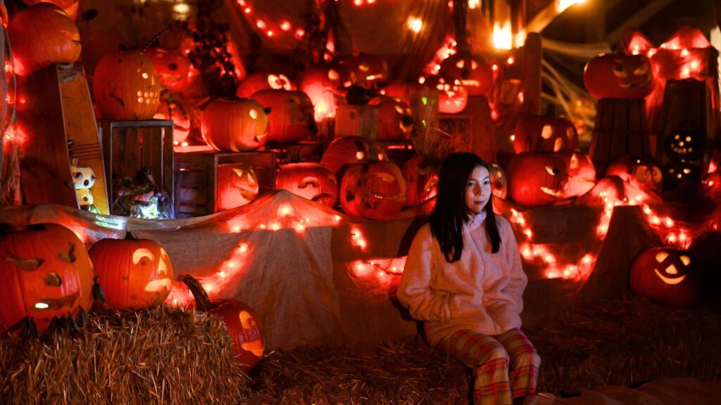 A person in a pink sweater sits on hay bales surrounded by glowing carved pumpkins and string lights in a festive Halloween display.