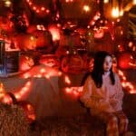 A person in a pink sweater sits on hay bales surrounded by glowing carved pumpkins and string lights in a festive Halloween display.
