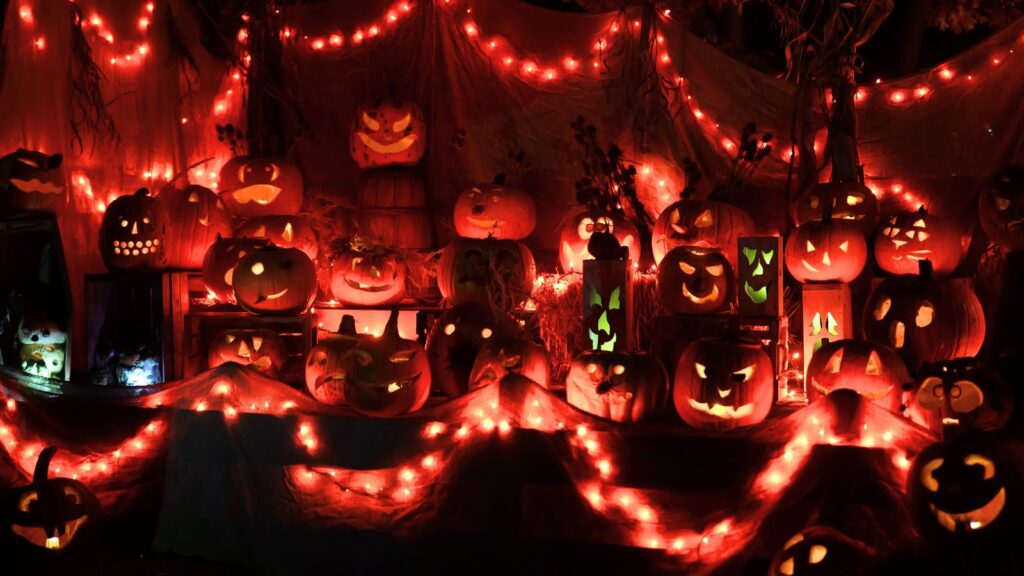 A display of carved jack-o’-lanterns is illuminated by orange and red string lights in a Halloween-themed nighttime setting.