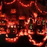 A display of carved jack-o’-lanterns is illuminated by orange and red string lights in a Halloween-themed nighttime setting.