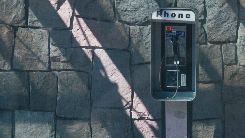 A payphone mounted on a stone wall, with sunlight casting shadows across the background, evokes the classic payphone booth designs America left behind.