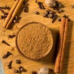 A bowl of ground brown spice is surrounded by cinnamon sticks, whole cloves, nutmeg, and allspice on a wooden surface.
