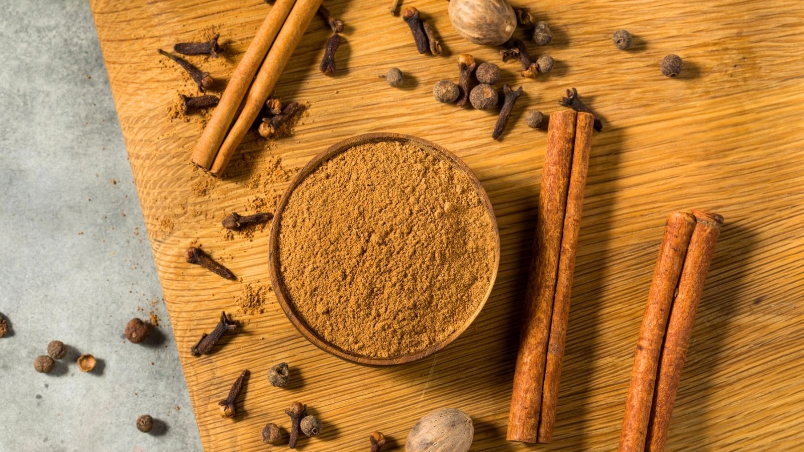 A bowl of ground brown spice is surrounded by cinnamon sticks, whole cloves, nutmeg, and allspice on a wooden surface.