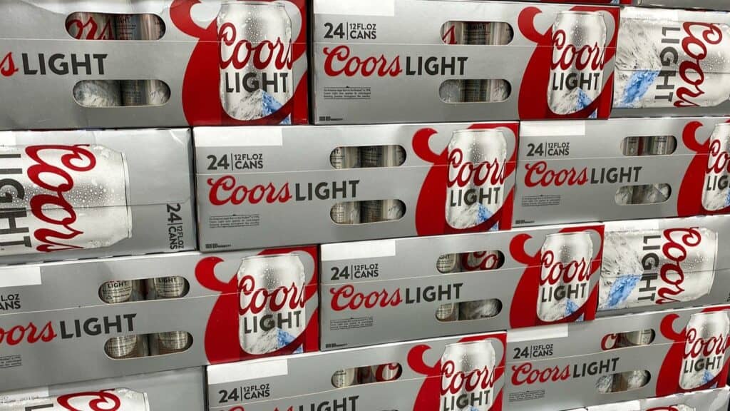 Stacks of 24-pack cases of Coors Light beer cans arranged on store shelves, featuring silver packaging with red and black text, evoke the vibrant NFL stadium beer culture that fans enjoy during game days.