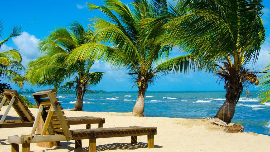 Wooden lounge chairs and palm trees on a sandy beach with blue ocean waves and a partly cloudy sky in the background.
