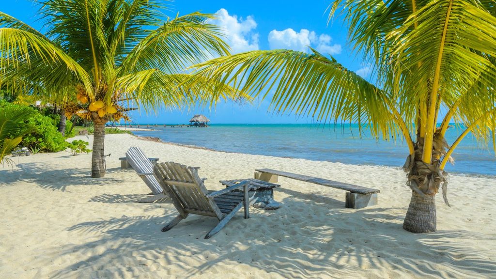 Two wooden chairs and a bench sit on a sandy beach under palm trees, facing calm turquoise water with a dock and thatched hut in the distance.