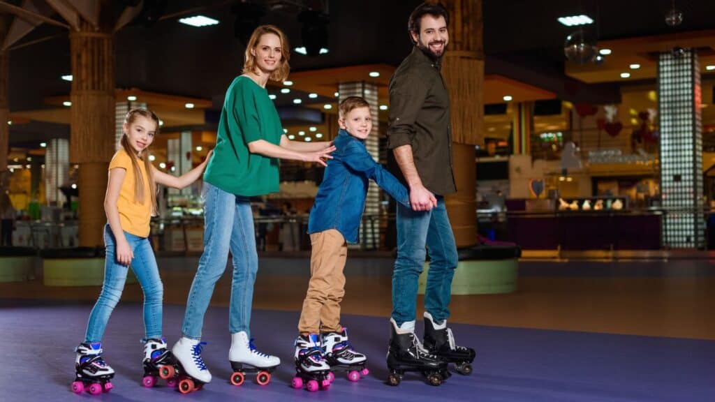 A family of four, two adults and two children, glide across the floor in a line while holding onto each other, embracing roller rink traditions as they enjoy roller skating indoors together.