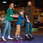 A family of four, two adults and two children, glide across the floor in a line while holding onto each other, embracing roller rink traditions as they enjoy roller skating indoors together.