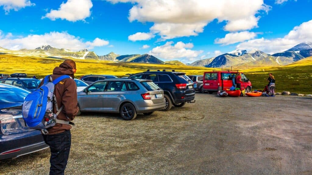 People stand near parked cars in a mountain parking lot under a blue sky with clouds and distant snow-capped peaks, enjoying the breathtaking scenery on one of the national park fee free days.