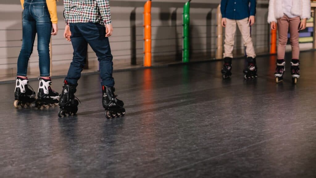 Four children wearing rollerblades stand on a roller rink indoors, facing each other. Only their legs and skates are visible, capturing a snapshot of timeless roller rink traditions.