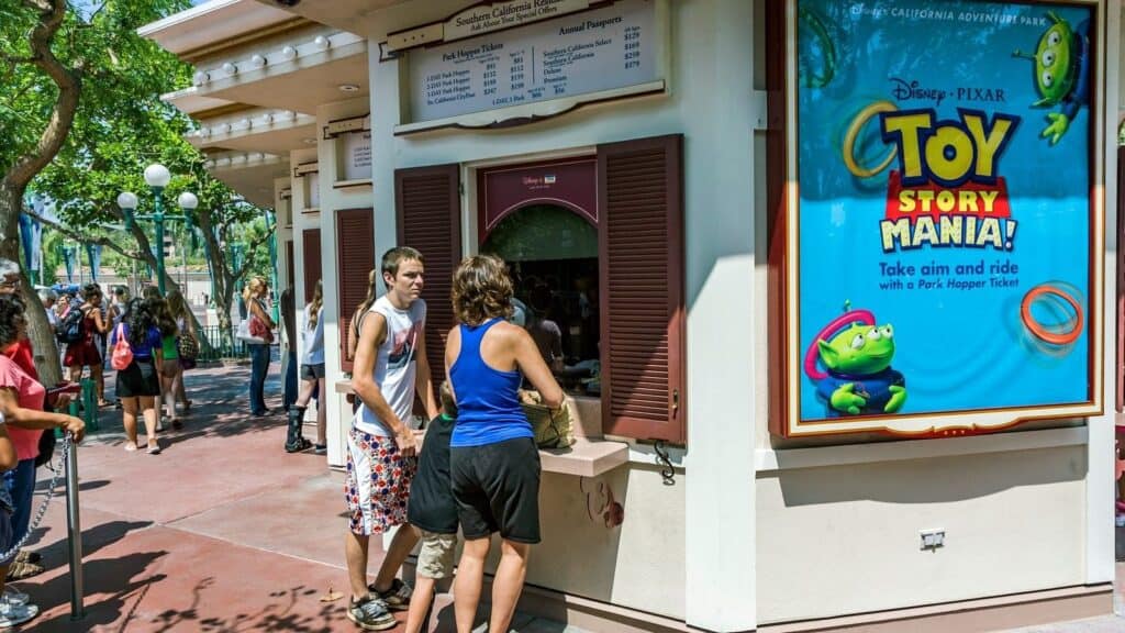 People purchase tickets at an outdoor booth near a large Toy Story Mania advertisement in a theme park setting, ready to enjoy the excitement while following Disney and Universal theme park rules for an unforgettable day.