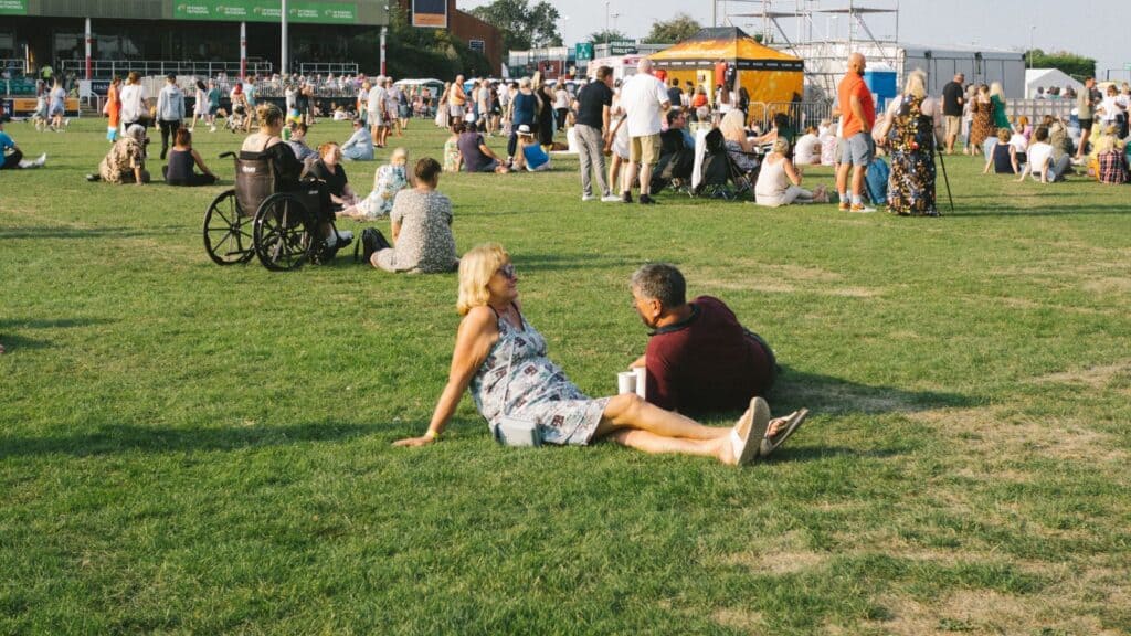 People sit and relax on a grassy field at an outdoor event, with groups scattered and some tents and buildings visible in the background under clear skies, capturing the spirit of America&rsquo;s greatest game day tradition.