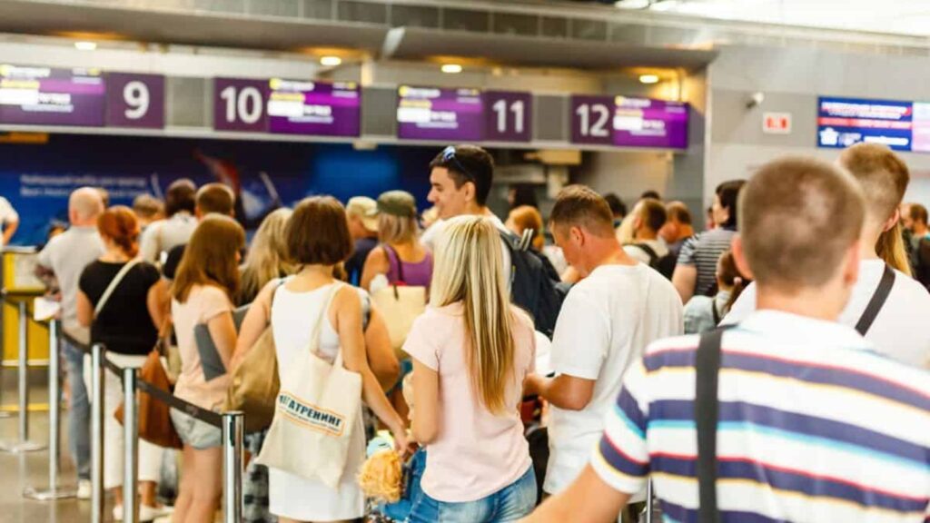 People wait in line at an airport check-in counter with numbered signs above, carrying bags and luggage.
