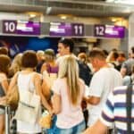 People wait in line at an airport check-in counter with numbered signs above, carrying bags and luggage.