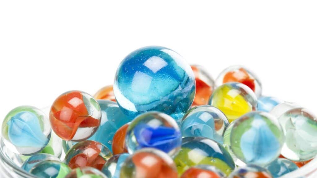 A variety of colorful glass marbles, with a large blue marble positioned on top of several smaller marbles, all against a white background—reminiscent of childhood days when board games disappeared from store shelves and simple toys like these brought hours of joy.