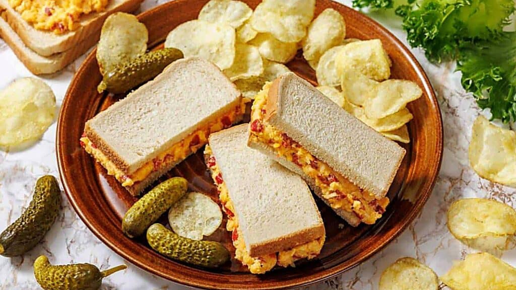 Three pimento cheese sandwiches on a brown plate with potato chips and pickles, surrounded by extra chips, pickles, and leafy greens.