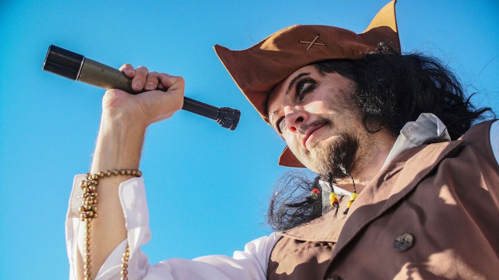 Person dressed as a pirate in brown clothing and hat holds a telescope up to their eye, looking into the distance against a clear blue sky, ready to celebrate Talk Like a Pirate Day with adventurous spirit.