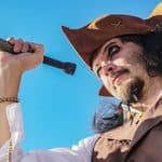 Person dressed as a pirate in brown clothing and hat holds a telescope up to their eye, looking into the distance against a clear blue sky, ready to celebrate Talk Like a Pirate Day with adventurous spirit.