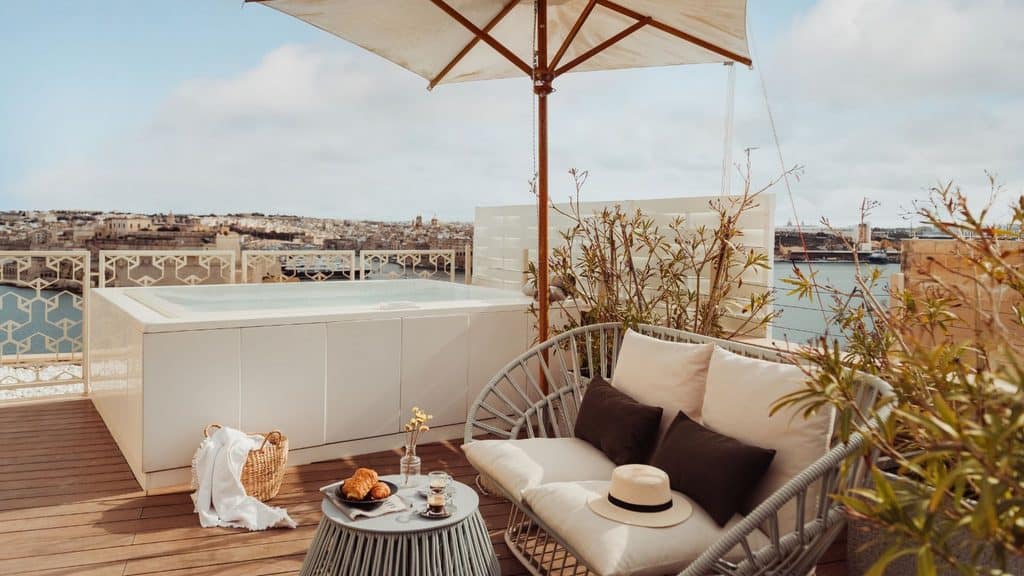 Outdoor terrace with a jacuzzi, two-seater sofa, small table with breakfast items, sun umbrella, and a city and harbor view in the background.