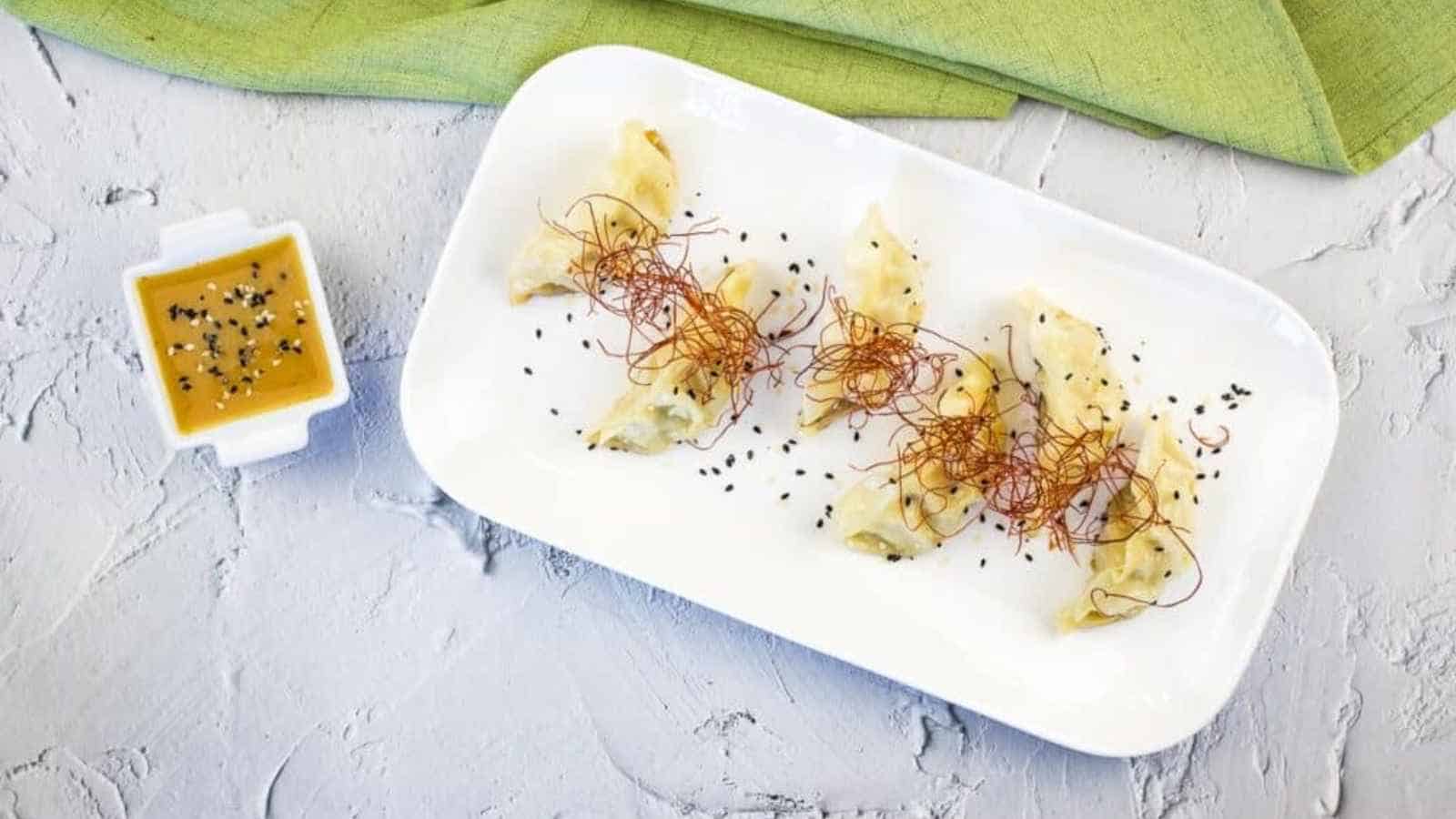 A white platter of pan-fried dumplings garnished with chili threads and black sesame seeds, served with a small bowl of dipping sauce.