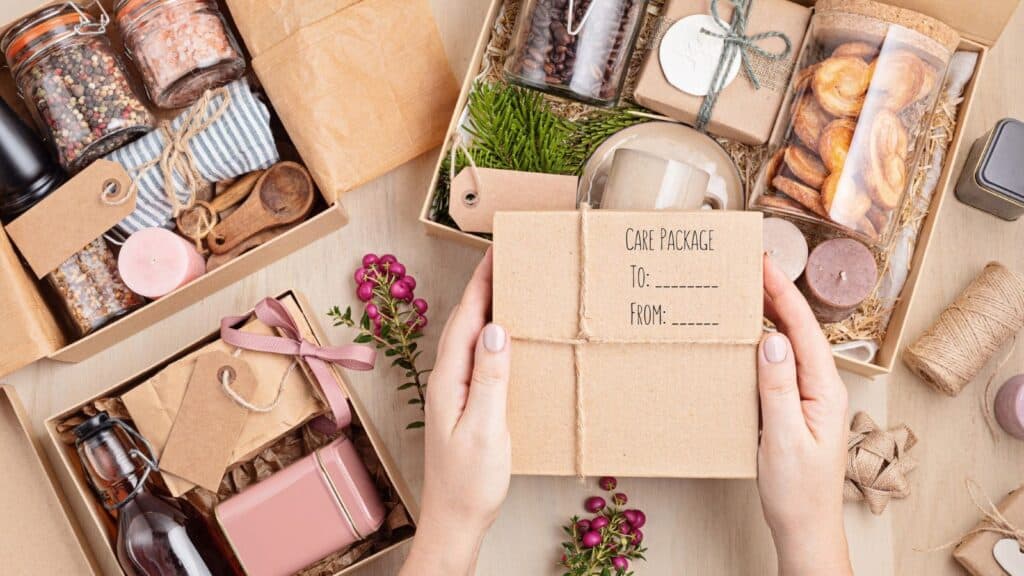 A person holds a care package box labeled “To” and “From,” surrounded by open gift boxes from College Care Package Co, displaying various items like candles, snacks, and bottles on a wooden table.