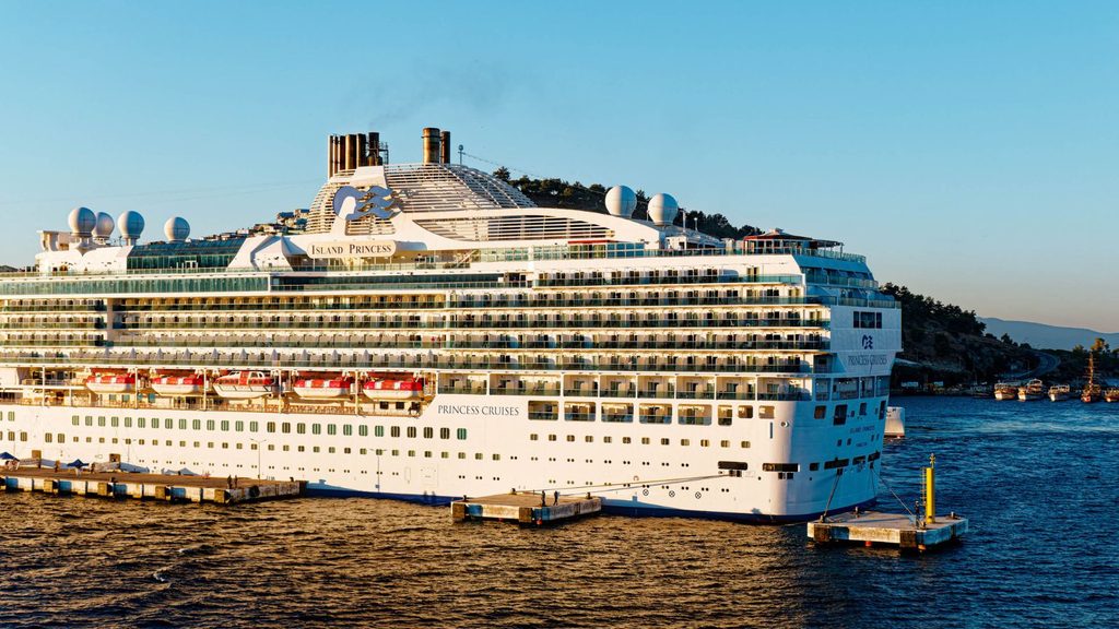 Large white cruise ship docked at a port, with calm blue water and hilly landscape in the background under a clear sky.