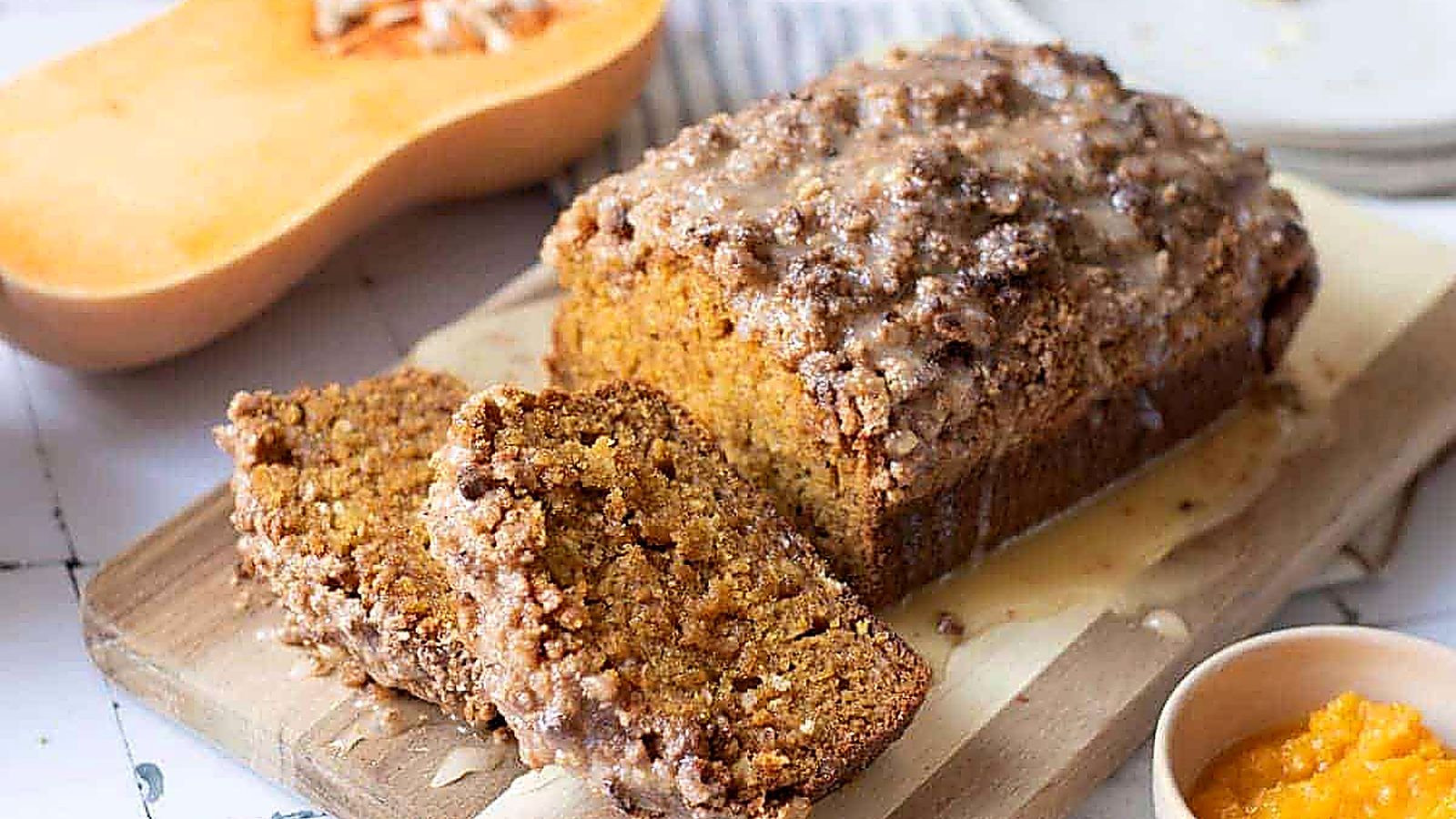 A loaf of Pumpkin Bread with Streusel & Glaze sits on a cutting board, with two slices cut. 