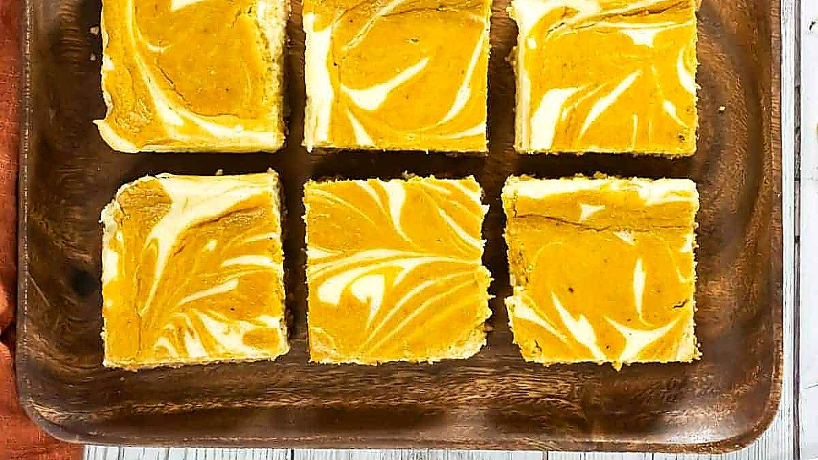 Six square pumpkin cheesecake bars are arranged on a wooden tray, displaying marbled orange and white patterns on top.