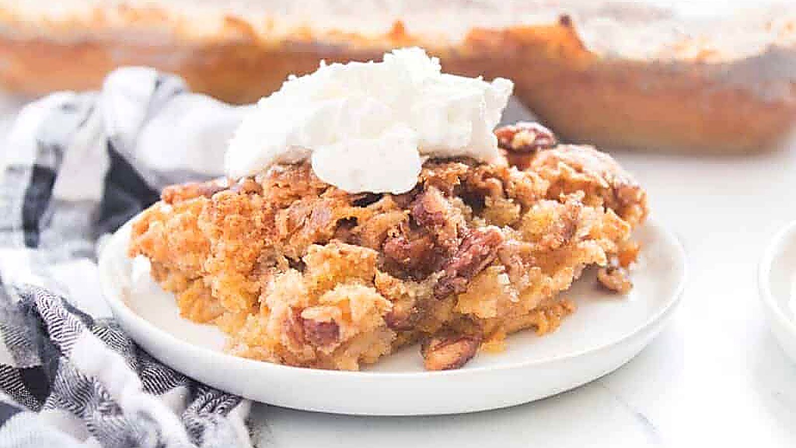 A slice of Pumpkin Crunch Cake served on a white plate with a dollop of whipped cream, napkin beside it, and a baking dish in the background.