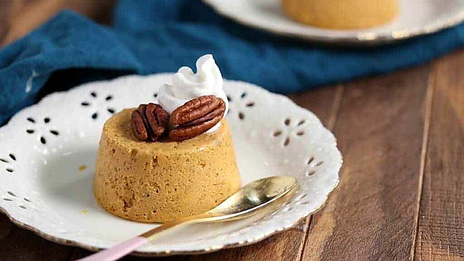 A small round pumpkin custard topped with pecans and whipped cream sits on a white plate with a gold spoon, on a wooden table.