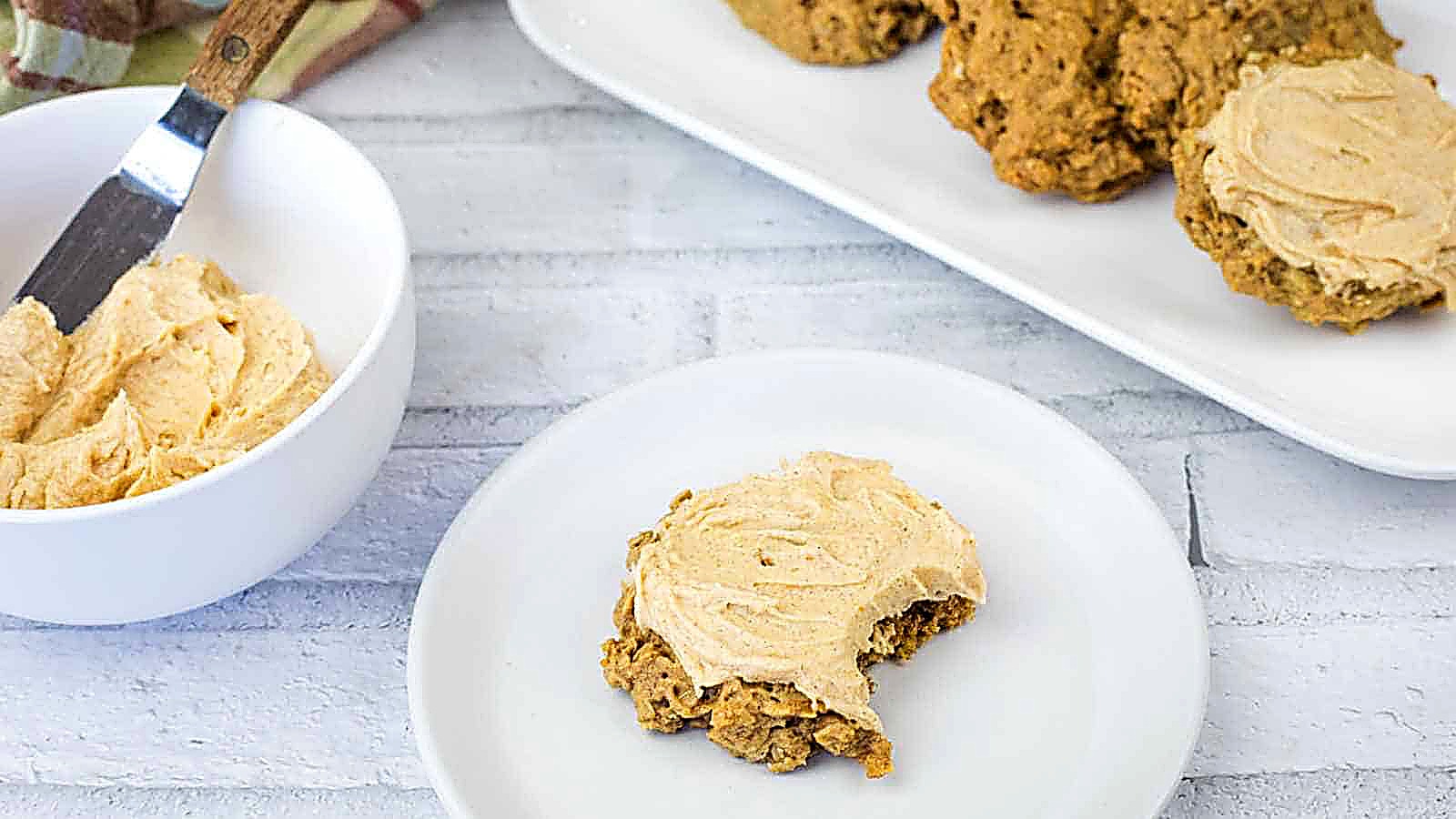 A pumpkin frosted cookie with a bite taken out sits on a plate. Nearby, a bowl of frosting with a knife and more cookies is on a platter.